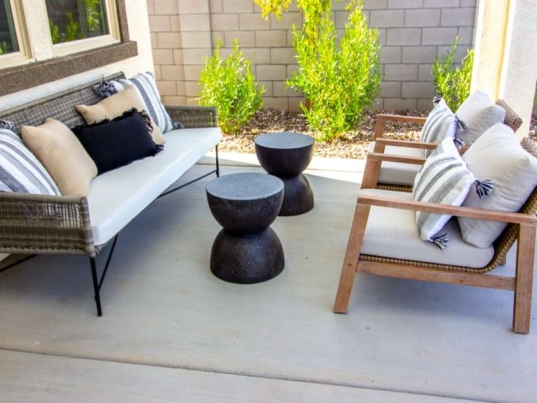 A concrete backyard patio with modern furniture. The furniture includes a couch, two chairs, and two small tables.
