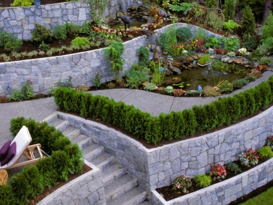 A backyard with landscape and hardscape features. The space has gray retaining walls, pathways, and greenery.