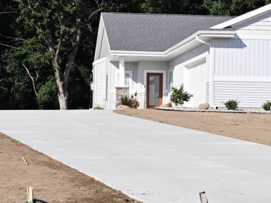 A light-colored concrete driveway near a white house. The house has a brown door and plants near the entrance.