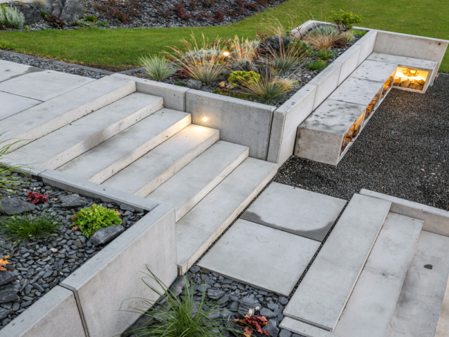 Light-gray colored concrete stairs and retaining walls surrounding landscape in a backyard by a grill.