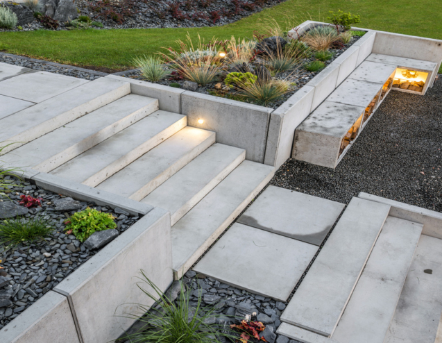 Light-gray colored concrete stairs and retaining walls surrounding landscape in a backyard by a grill.