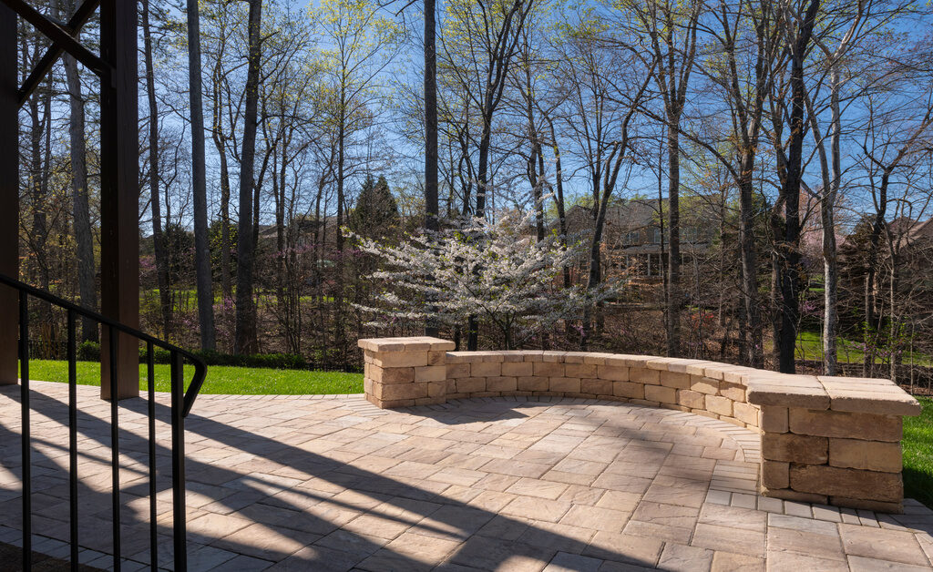 A rounded multi-colored stone wall at the end of a patio in a backyard. Grass and trees fill in the background.
