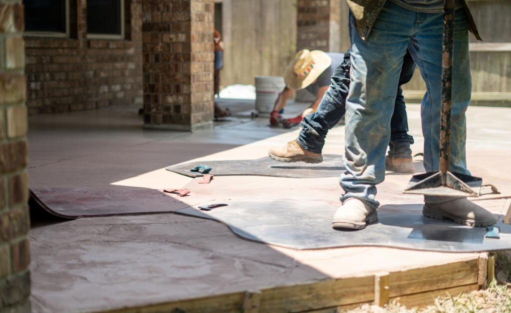 Four contractors installing a large colored concrete patio in a residential backyard on a sunny day.