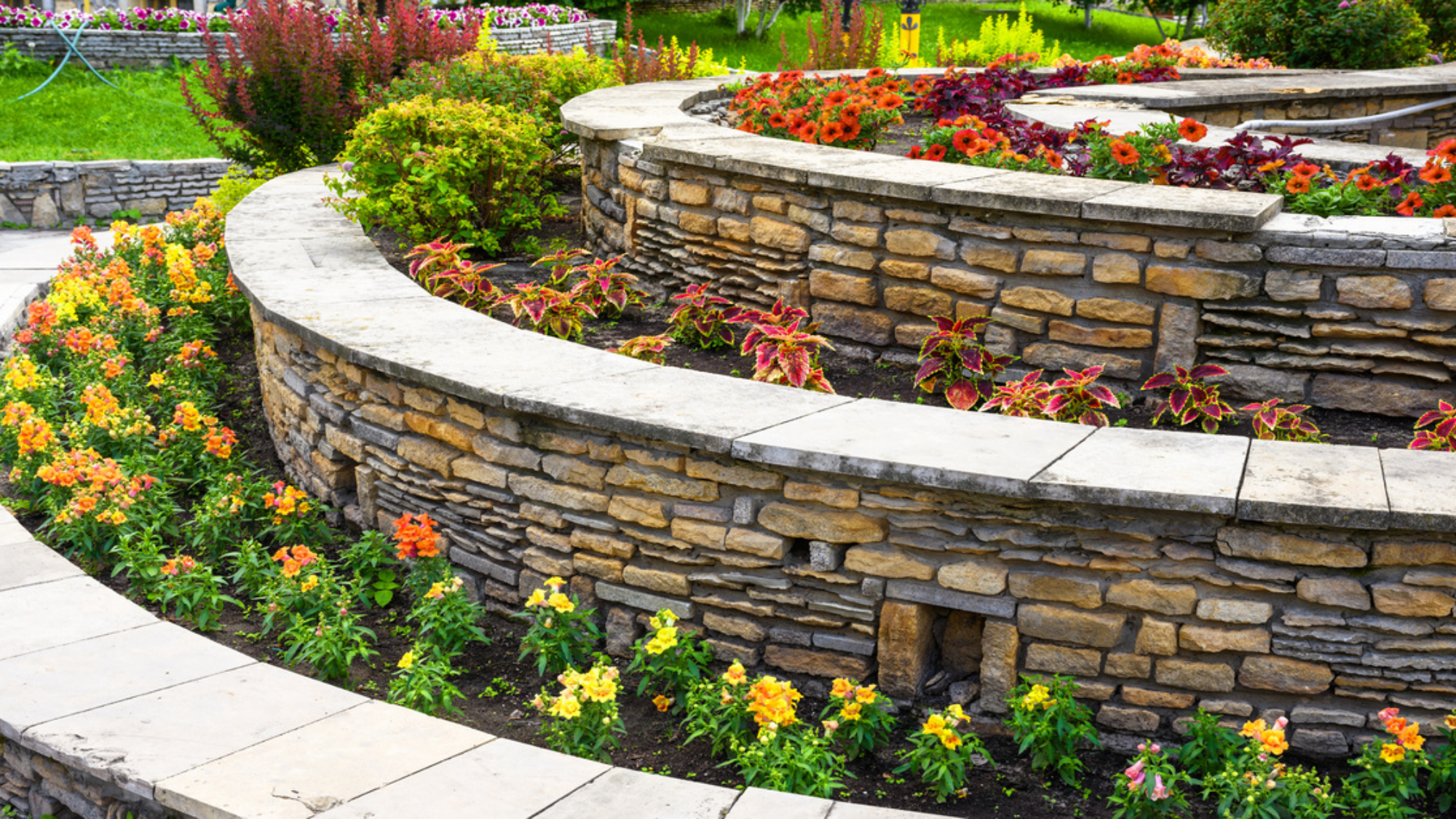 A terraced garden contains curved stone walls, colorful flowers, lush greenery, and a light paved walkway.