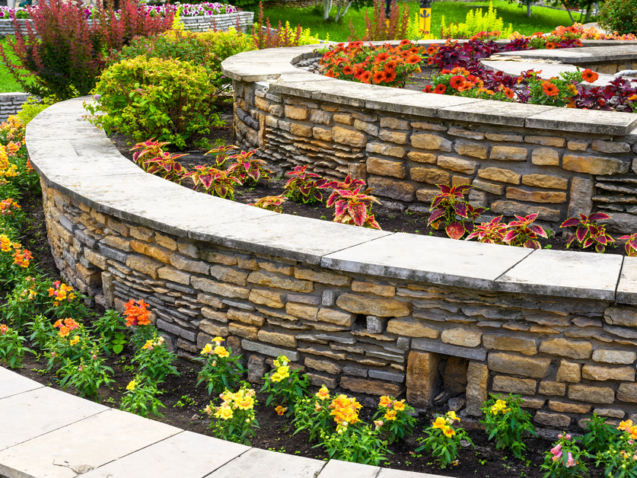 A terraced garden contains curved stone walls, colorful flowers, lush greenery, and a light paved walkway.
