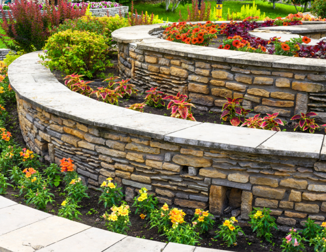A terraced garden contains curved stone walls, colorful flowers, lush greenery, and a light paved walkway.