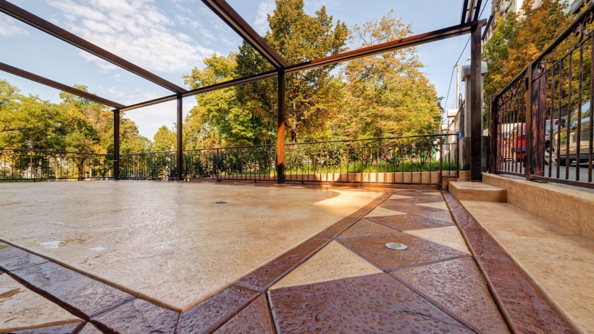 A stamped concrete patio with geometric patterns sits beneath open pergola frame, bordered by iron fence and fall trees.