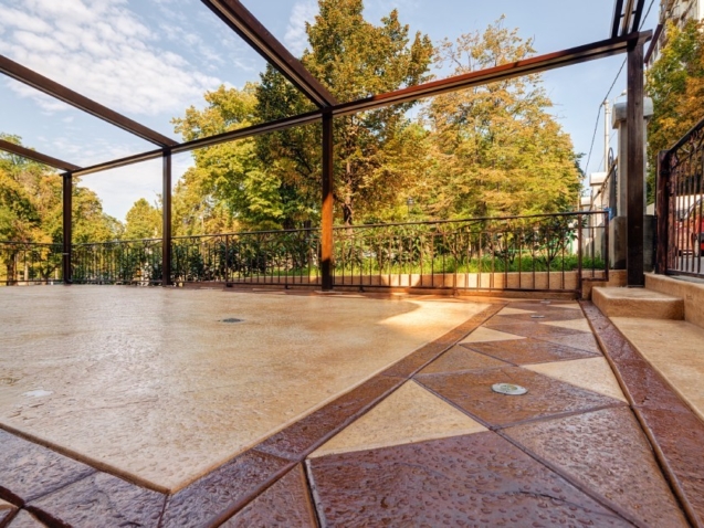 A stamped concrete patio with geometric patterns sits beneath open pergola frame, bordered by iron fence and fall trees.