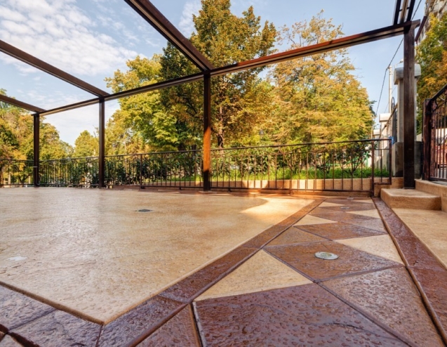 A stamped concrete patio with geometric patterns sits beneath open pergola frame, bordered by iron fence and fall trees.