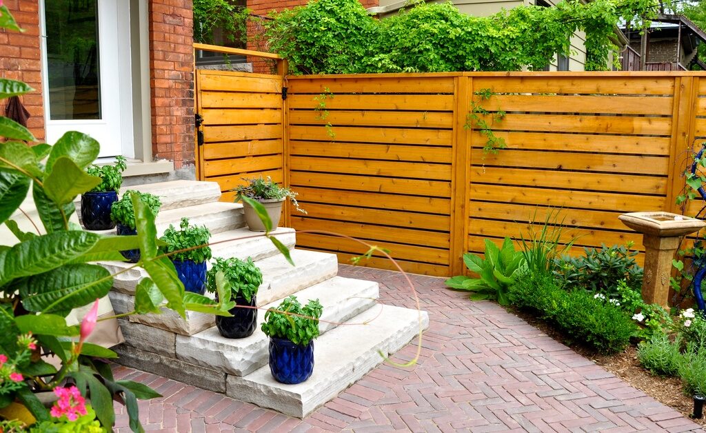 A brick paver patio features stone steps with potted plants beside a wooden privacy fence in a landscaped backyard.