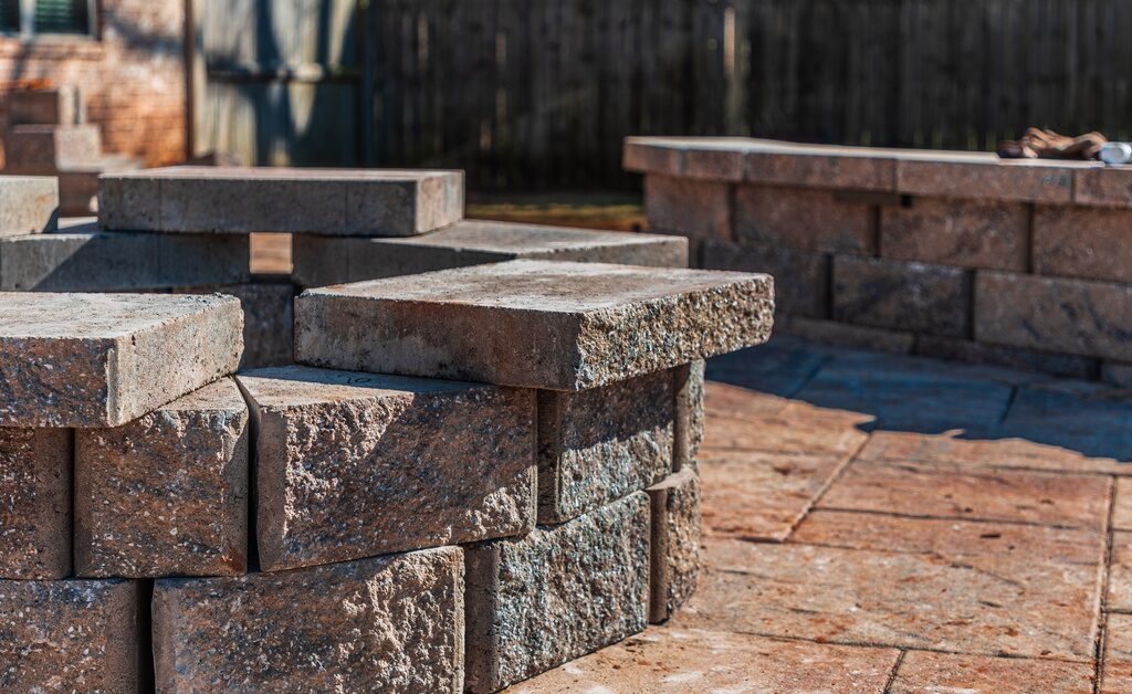 A stone fire pit under construction sits on a paver patio in a backyard with retaining walls visible behind it.