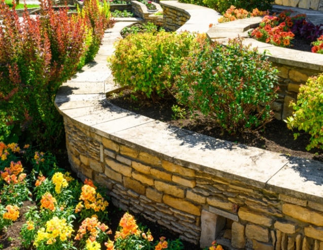 A tiered stone residential retaining wall with colorful shrubs and blooming flowers supports a curved walkway.