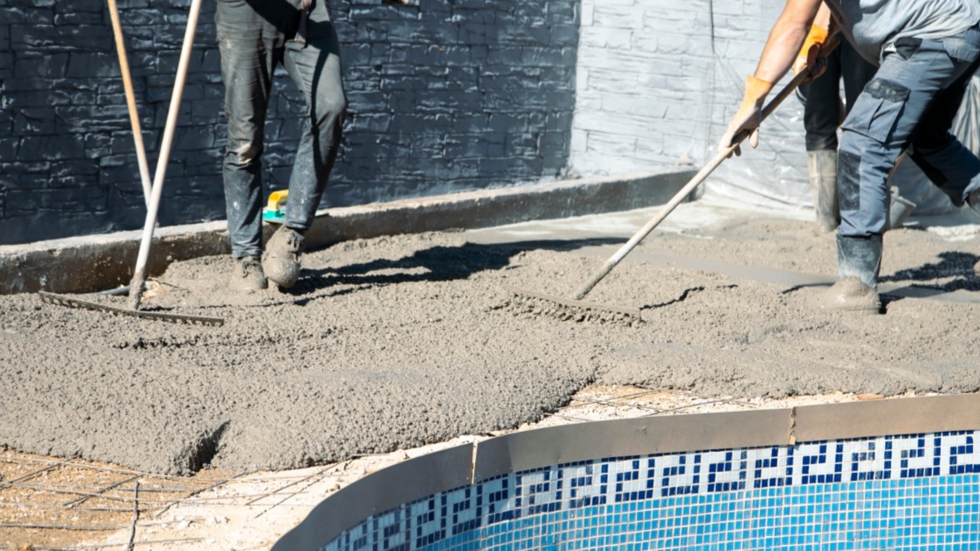Workers wearing boots spread wet concrete with rakes along the edge of a pool on a bright sunny day.