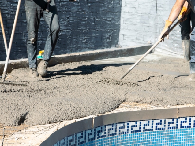 Workers wearing boots spread wet concrete with rakes along the edge of a pool on a bright sunny day.