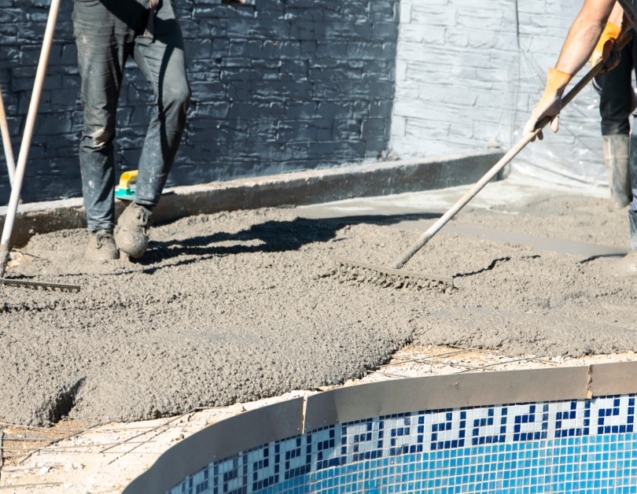 Workers wearing boots spread wet concrete with rakes along the edge of a pool on a bright sunny day.