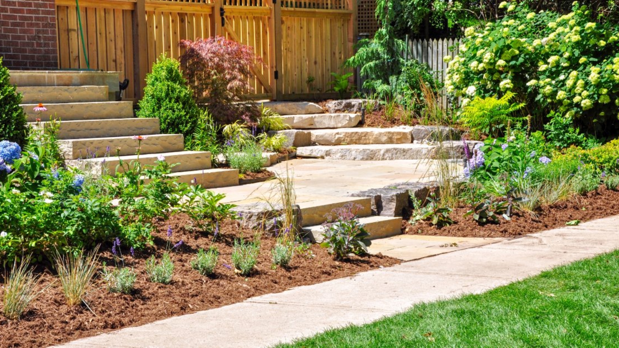 A stone stairway leads through a lush garden beside a wooden fence, brick wall, and bright green lawn.
