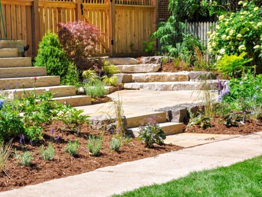 A stone stairway leads through a lush garden beside a wooden fence, brick wall, and bright green lawn.