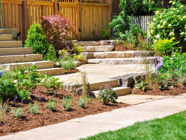 A stone stairway leads through a lush garden beside a wooden fence, brick wall, and bright green lawn.