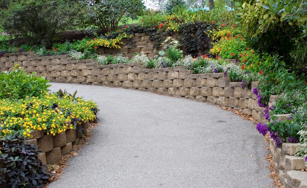 A block retaining wall lines a curved driveway beside layered flower beds and dense green shrubs on both sides.