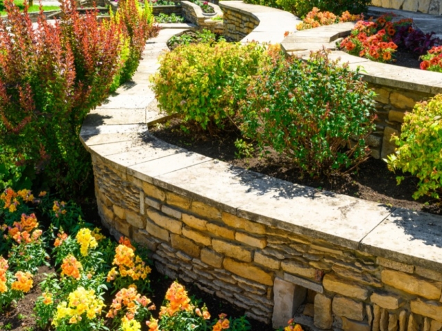 A stone retaining wall curves around bright flower beds and trimmed shrubs in a sunny tiered garden.