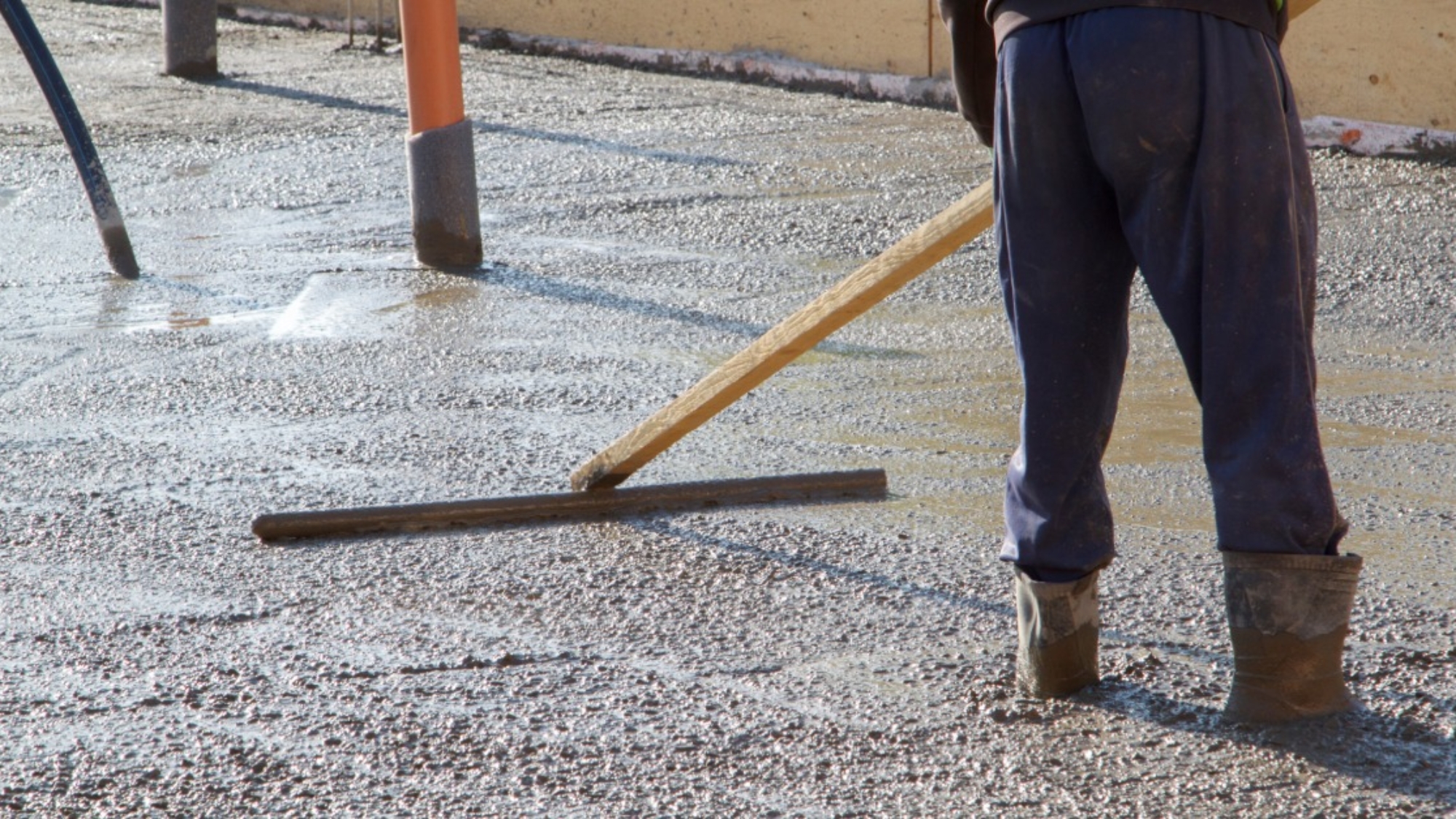 A worker levels a broad slab of wet concrete with a long screed while standing inside the framed pour area.