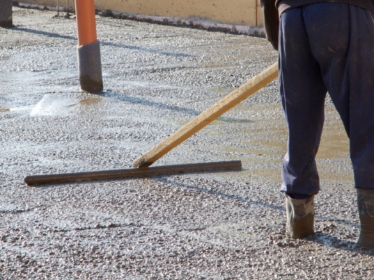 A worker levels a broad slab of wet concrete with a long screed while standing inside the framed pour area.
