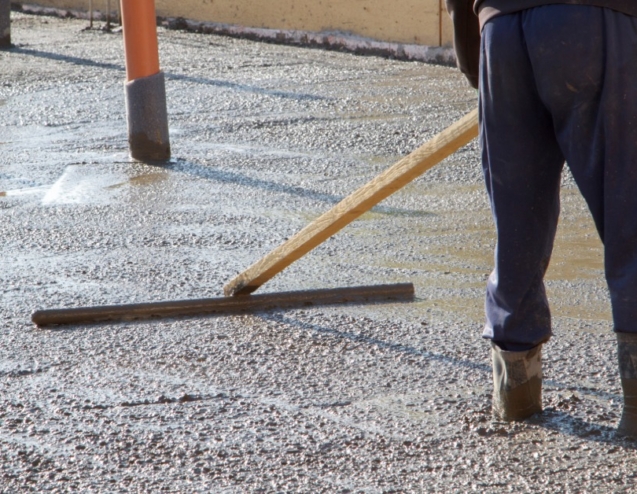A worker levels a broad slab of wet concrete with a long screed while standing inside the framed pour area.