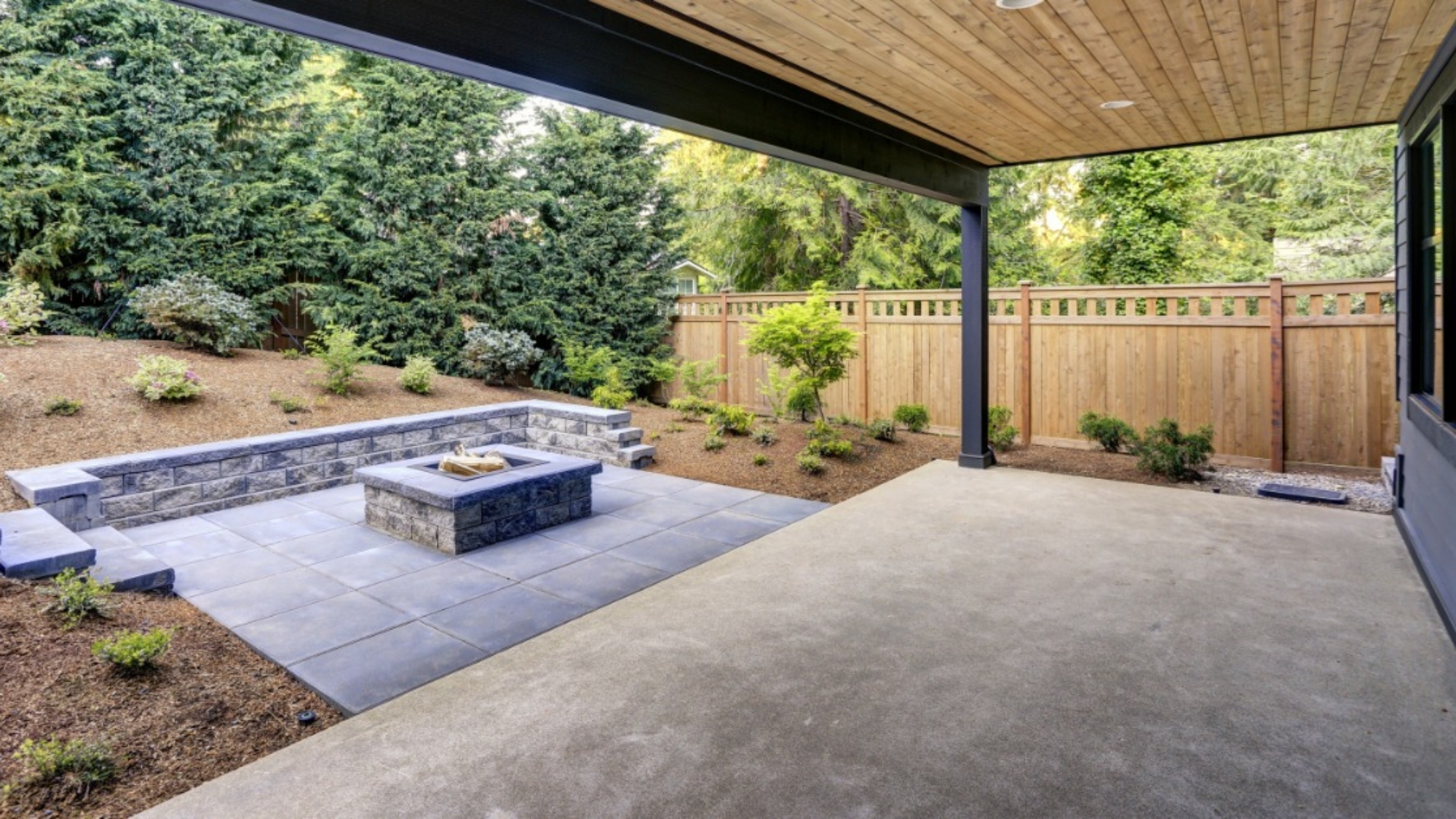 A covered backyard patio combines a smooth concrete slab with gray pavers, a fire pit, and a landscaped garden.
