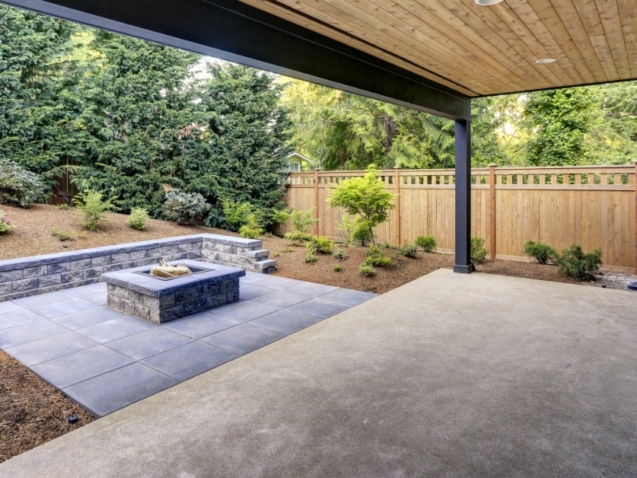 A covered backyard patio combines a smooth concrete slab with gray pavers, a fire pit, and a landscaped garden.