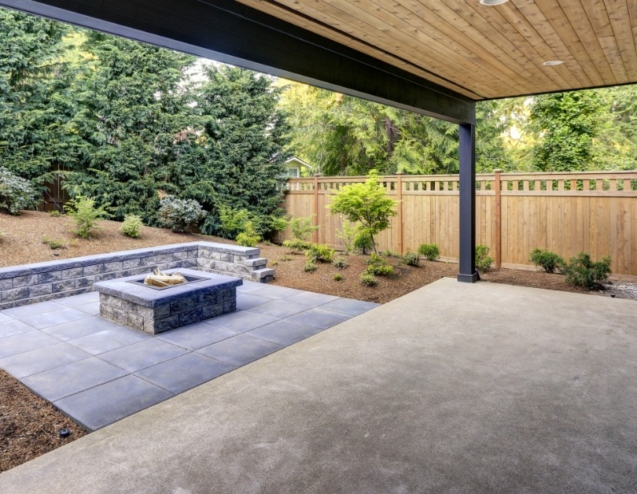 A covered backyard patio combines a smooth concrete slab with gray pavers, a fire pit, and a landscaped garden.