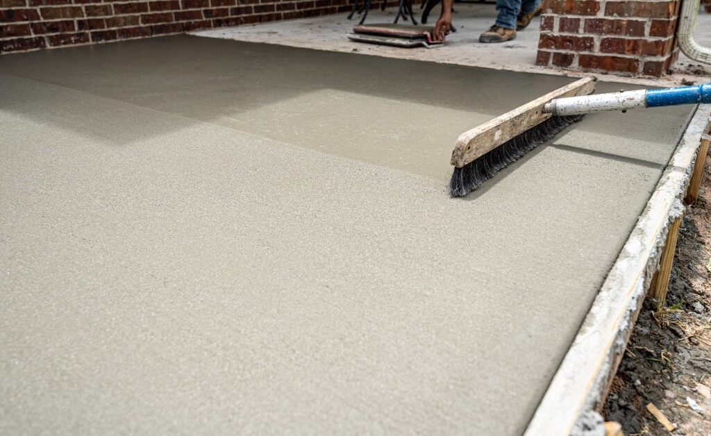 A concrete contractor uses a broom to texture a freshly poured concrete patio beside a red brick home.