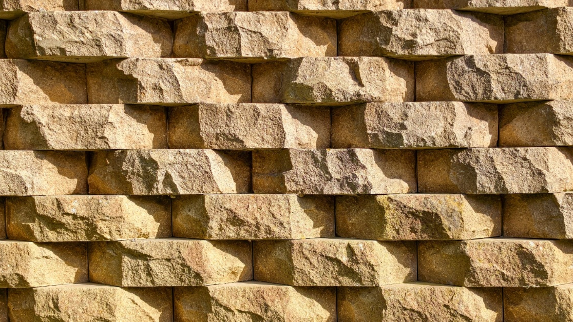 Rows of tan retaining wall blocks featuring rough, stone-like faces with warm color variations and deep shadowed joints.