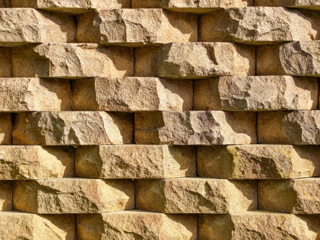 Rows of tan retaining wall blocks featuring rough, stone-like faces with warm color variations and deep shadowed joints.