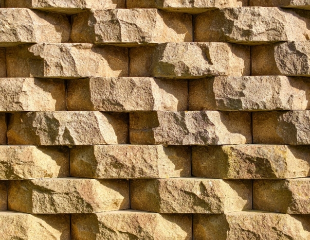 Rows of tan retaining wall blocks featuring rough, stone-like faces with warm color variations and deep shadowed joints.