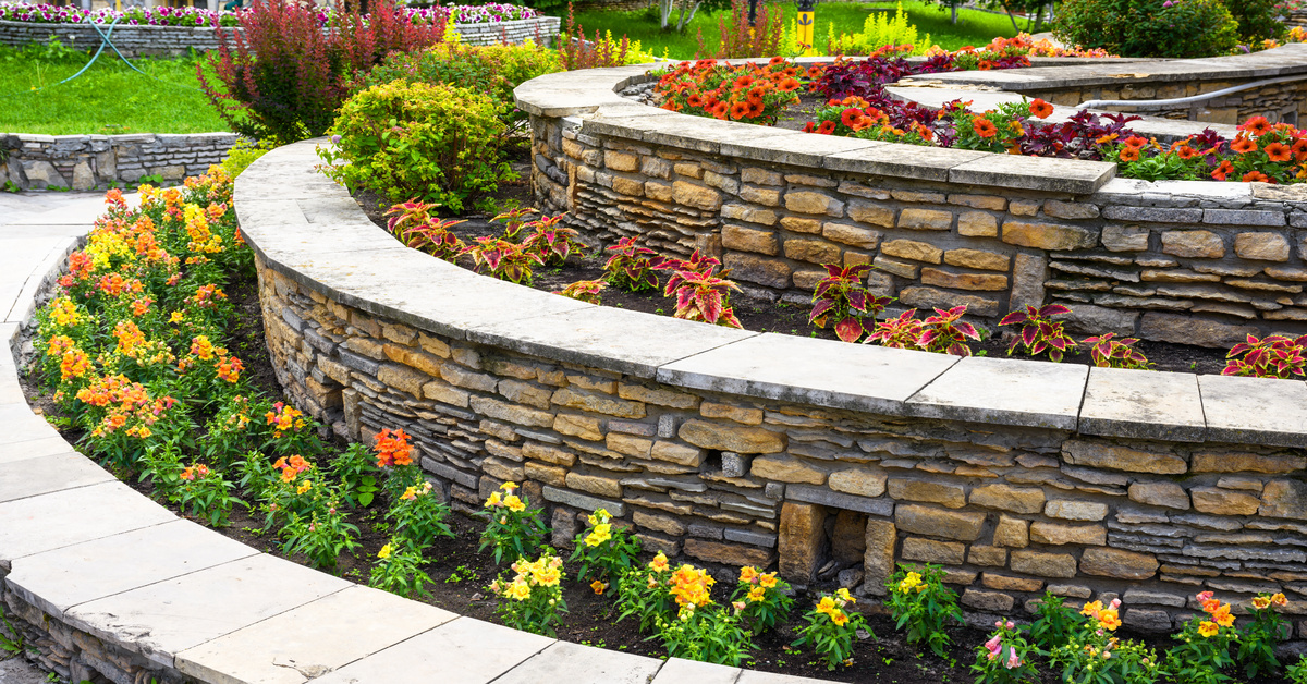A terraced garden contains curved stone walls, colorful flowers, lush greenery, and a light paved walkway.
