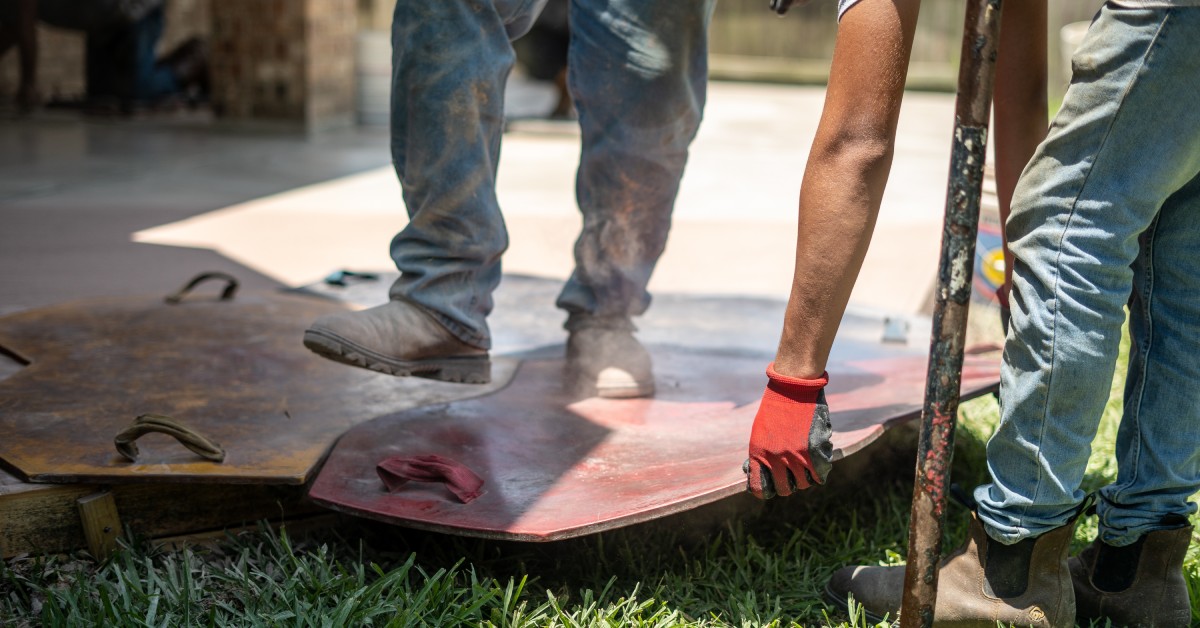 Workers wearing boots and gloves lift concrete stamping mats as they imprint a fresh patio slab on a sunny day.