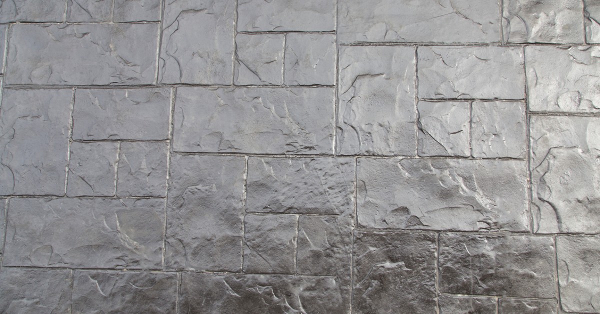 A stamped concrete patio features a rectangular stone pattern with textured gray finish, showing realistic joints and depth.