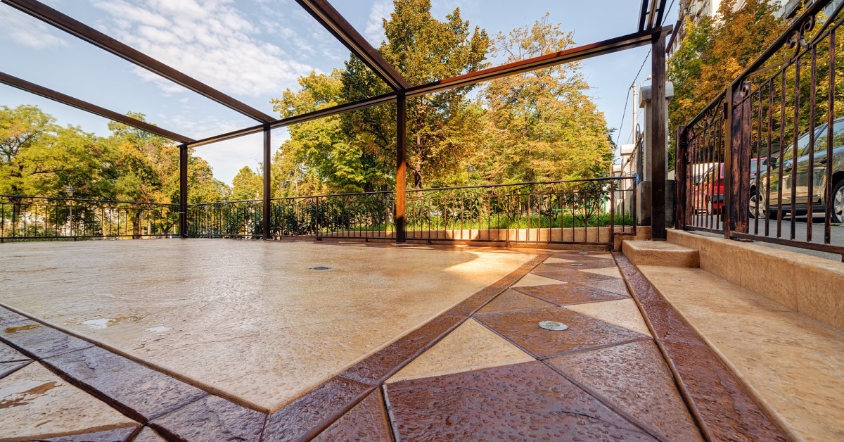 A stamped concrete patio with geometric patterns sits beneath open pergola frame, bordered by iron fence and fall trees.