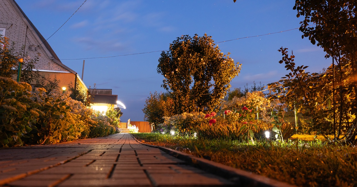 A brick paver walkway leads through an illuminated backyard garden at dusk with trees and landscape lighting visible.