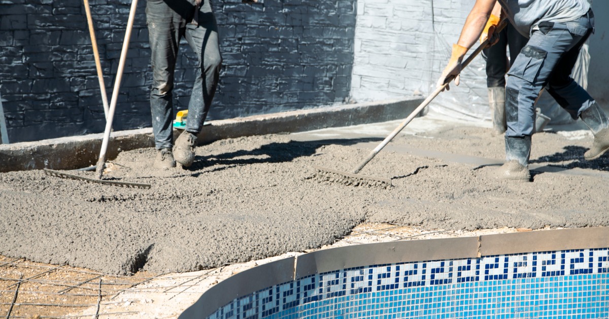 Workers wearing boots spread wet concrete with rakes along the edge of a pool on a bright sunny day.