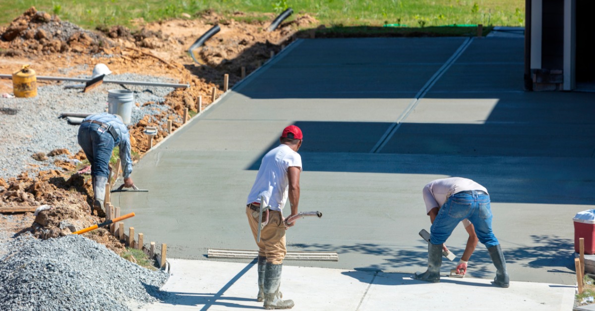 Three workers wearing boots smooth and finish a large concrete driveway near a new home on a bright sunny day.