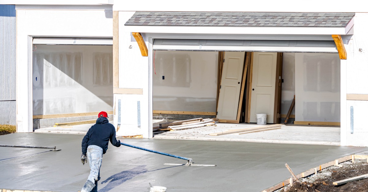 A worker smooths a freshly poured concrete driveway in front of a new house with two open garage doors.