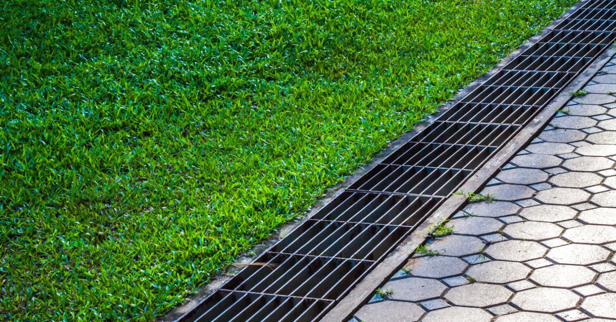 A metal trench drain channels water between a green lawn and a paved walkway with hexagonal stone pavers.