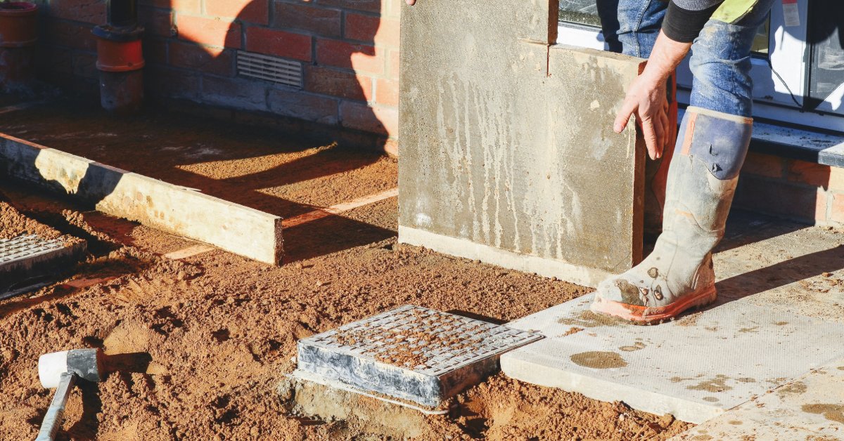 A worker wearing boots lifts a concrete slab beside a brick home while wet soil and utility covers sit on the ground nearby.