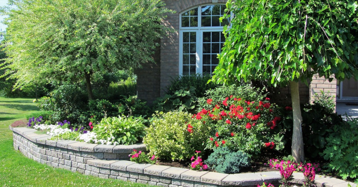 A low block retaining wall frames colorful garden beds beside a brick home and a manicured front lawn.