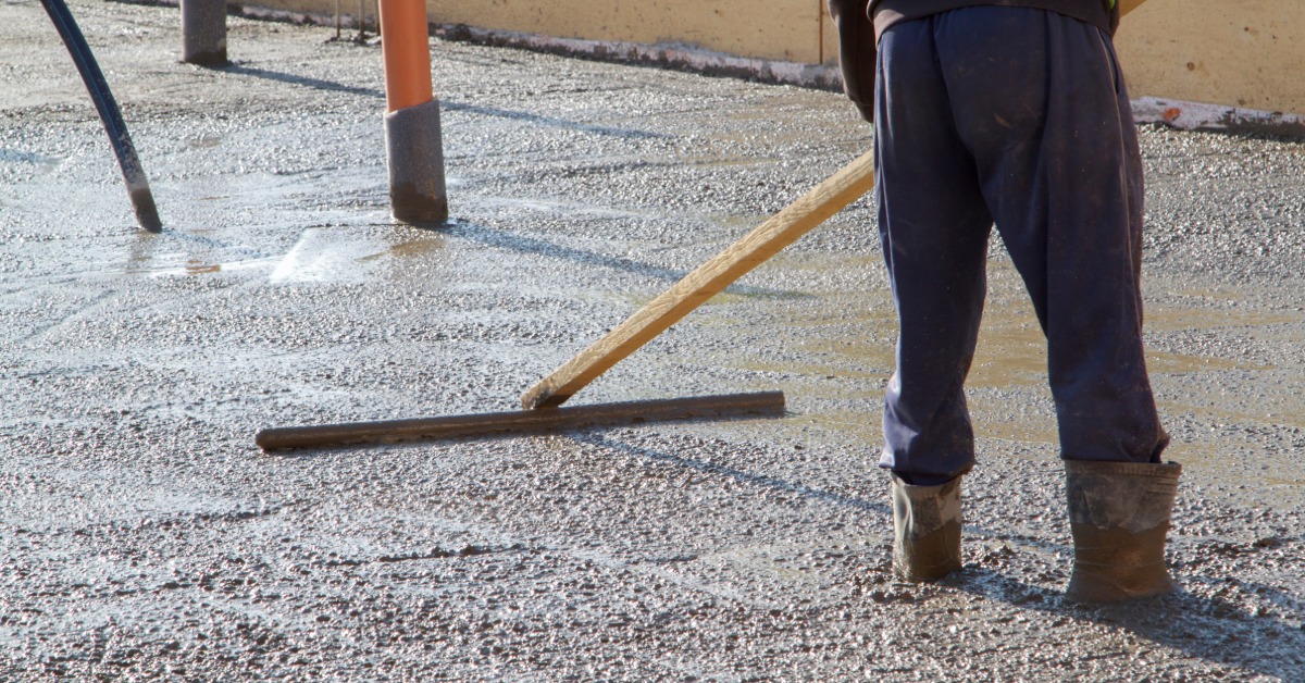 A worker levels a broad slab of wet concrete with a long screed while standing inside the framed pour area.