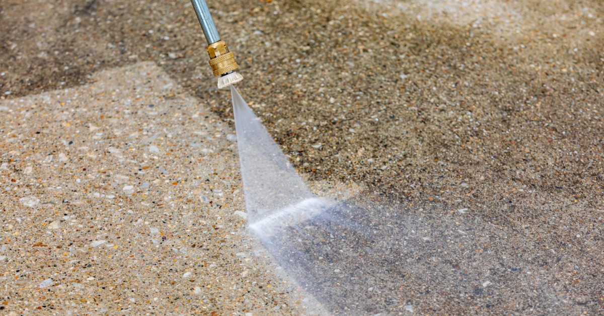 A pressure washer sprays a concrete surface, lifting dark grime to reveal a lighter, cleaner section below.