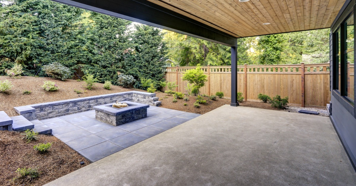 A covered backyard patio combines a smooth concrete slab with gray pavers, a fire pit, and a landscaped garden.