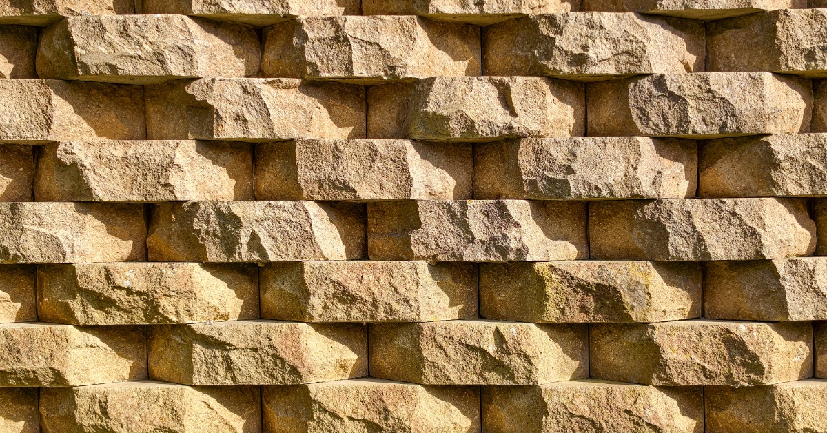 Rows of tan retaining wall blocks featuring rough, stone-like faces with warm color variations and deep shadowed joints.